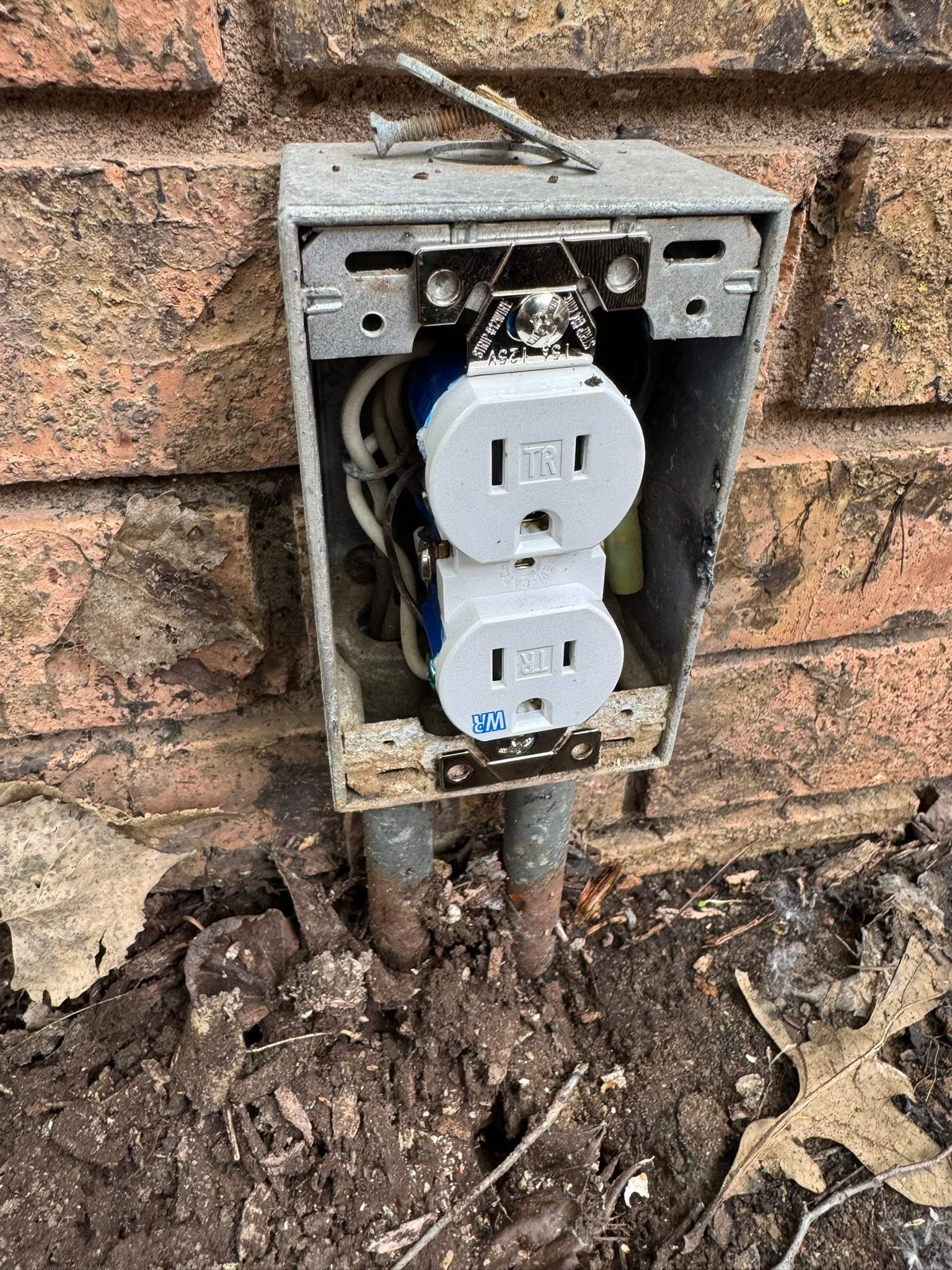 Open outdoor electrical outlet box with exposed wiring on a brick wall near muddy ground